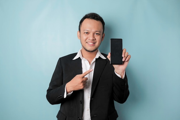 Um retrato de um empresário asiático feliz está sorrindo e segurando seu smartphone mostrando espaço de cópia na tela do 39s vestindo terno preto isolado por um fundo azul