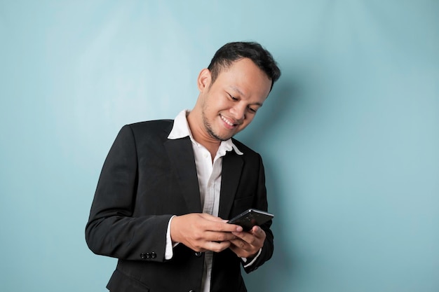 Um retrato de um empresário asiático feliz está sorrindo e segurando seu smartphone vestindo terno preto isolado por um fundo azul