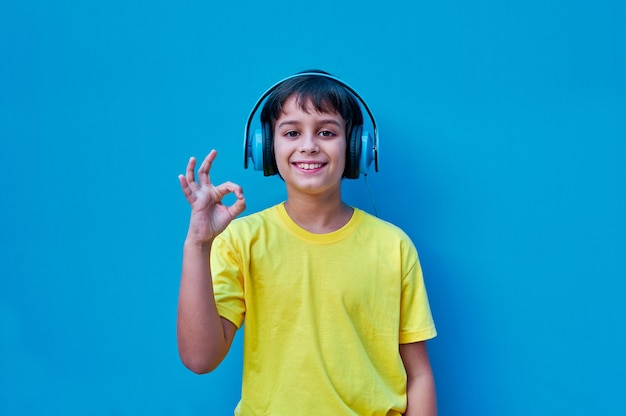 Um retrato de um menino sorridente com uma camiseta amarela e fones de ouvido azuis, fazendo um gesto de ok com a mão