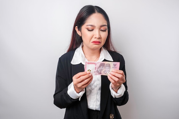 Um retrato de uma empresária asiática vestindo um terno preto e segurando dinheiro em rúpia indonésia isolado por fundo branco parece deprimido