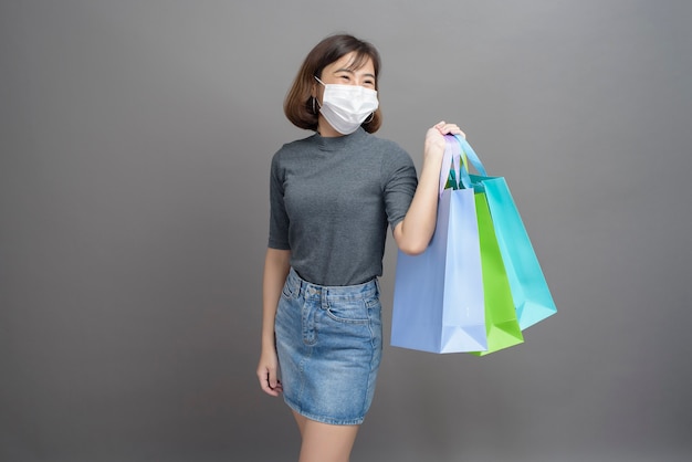 Um retrato de uma jovem e bela mulher asiática vestindo uma mak cirúrgica está segurando um cartão de crédito e uma sacola de compras colorida isolada em cinza