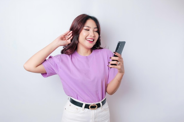 Um retrato de uma mulher asiática feliz está sorrindo e segurando seu smartphone usando uma camiseta roxa lilás isolada por um fundo branco