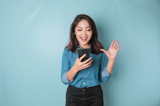 Um retrato de uma mulher asiática feliz está sorrindo e segurando seu smartphone vestindo uma camisa azul isolada por um fundo azul