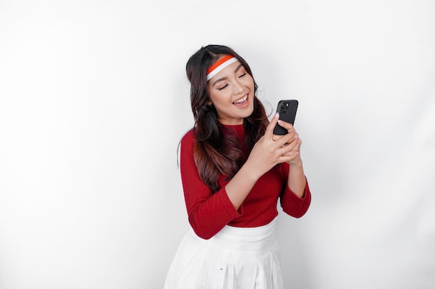 Um retrato de uma mulher asiática sorridente usando bandana e segurando seu telefone isolado pelo conceito de dia da independência da Indonésia de fundo branco
