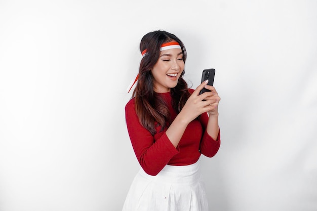 Um retrato de uma mulher asiática sorridente usando bandana e segurando seu telefone isolado pelo conceito de dia da independência da Indonésia de fundo branco