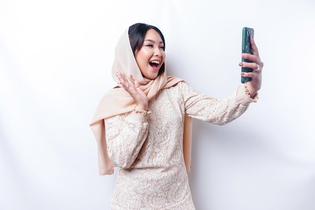 Um retrato de uma mulher muçulmana asiática feliz vestindo um hijab segurando seu telefone isolado por fundo branco