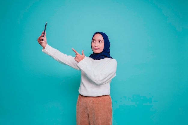 Um retrato de uma mulher muçulmana asiática feliz vestindo uma camisa branca e hijab segurando seu telefone isolado por fundo azul