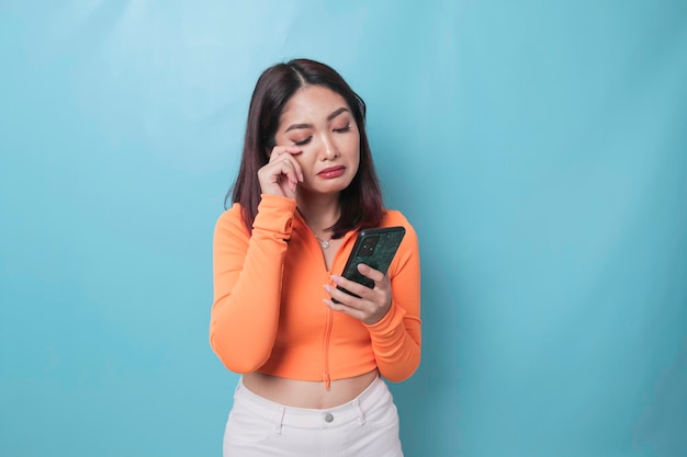 Um retrato de uma triste mulher asiática olhando para seu smartphone sobre fundo azul