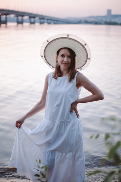 Um retrato gentil de uma encantadora mulher caucasiana em um vestido branco de verão e um chapéu de palha ...