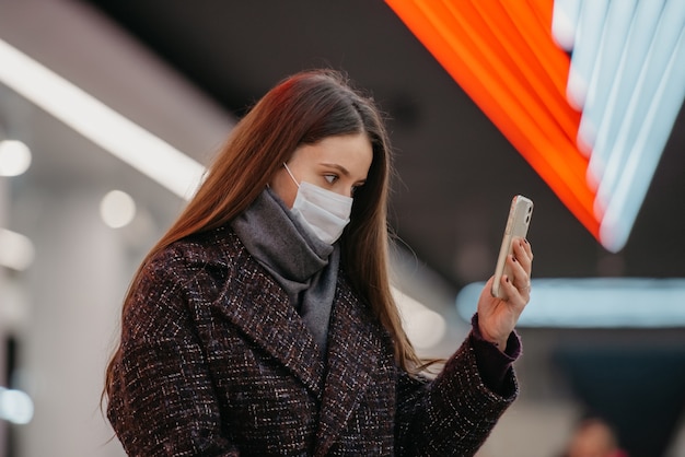 Um retrato próximo de uma mulher com uma máscara facial médica está sentada na estação de metrô com um smartphone e fazendo uma selfie. Uma garota com máscara cirúrgica está mantendo distância social no metrô