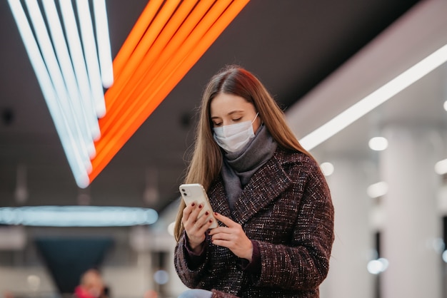Um retrato próximo de uma mulher com uma máscara facial médica está sentada na estação de metrô com um smartphone e lendo as notícias. Uma garota com máscara cirúrgica mantém distância social no metrô.