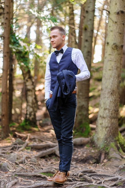 Um retrato sério de um noivo bonito em um terno azul e gravata borboleta está de pé contra o pano de fundo de vegetação na floresta.