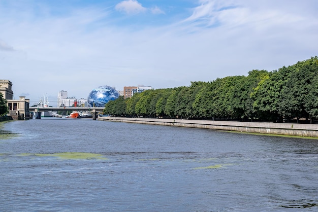 Um rio na parte turística da cidade com margens reforçadas com cercas de concreto e um parque com copas de árvores verdejantes