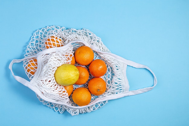 Um saco de cordas de algodão branco com tangerinas e limão encontra-se em um fundo azul