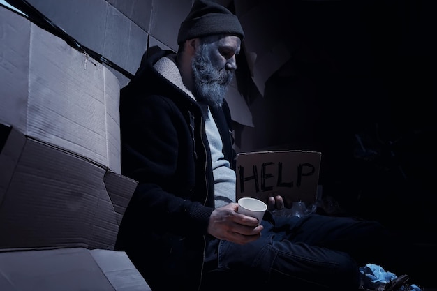 Um sem-teto barbudo senta-se em caixas na rua e pede ajuda. Precisa de um sem-teto pede dinheiro para comida e pernoite.