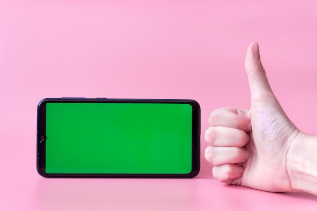 Um smartphone na posição horizontal com uma tela verde em um fundo rosa e uma mão com o polegar para cima, chroma key, mock up.