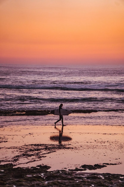 Um surfista caminha na praia com um céu rosa e laranja ao fundo.