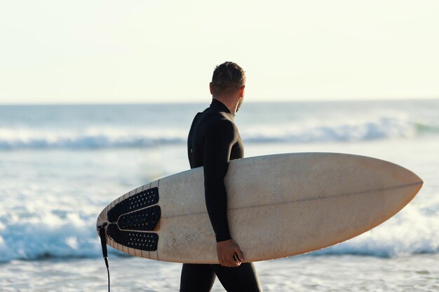 Um surfista em uma roupa de mergulho andando na praia segurando uma prancha e olhando para o mar