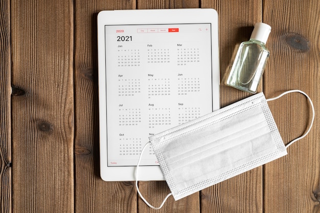 Um tablet com um calendário aberto para 2021 anos e máscara médica protetora e desinfetante para as mãos em um fundo de mesa de tábuas de madeira. conceito de proteção contra coronavírus covid-19 em 2021.