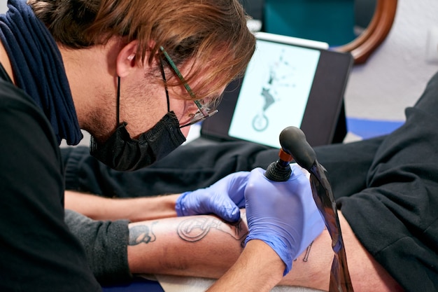 Um tatuador com luvas azuis olhando para um tablet e fazendo uma tatuagem na perna de um homem em um estúdio