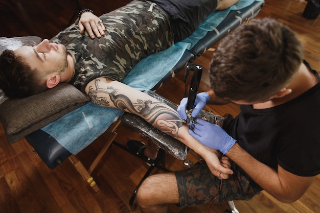 Um tatuador profissional fazendo uma tatuagem no braço de um jovem por uma máquina com tinta preta