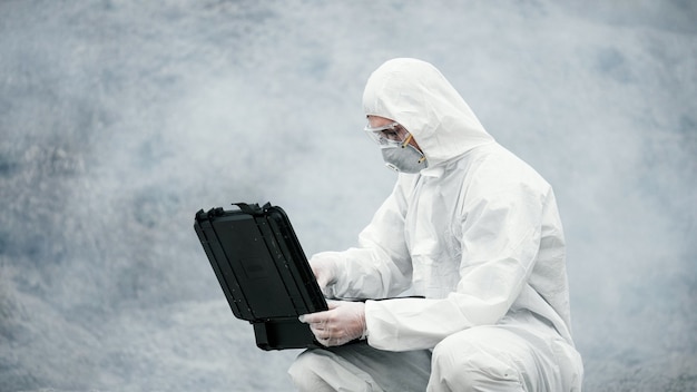 Um técnico de laboratório com uma máscara e uma roupa de proteção contra produtos químicos abre uma caixa de ferramentas em terra firme ao redor ...