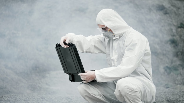 Um técnico de laboratório com uma máscara e uma roupa de proteção química abre uma caixa de ferramentas em terra firme