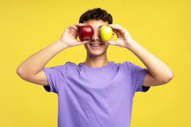 Um tipo engraçado e bonito com aparelhos ortopédicos a brincar com maçãs isoladas em fundo amarelo.