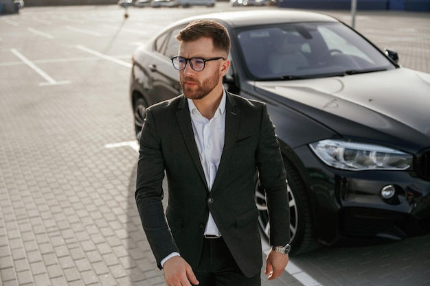 Foto um tipo esperto e bonito com óculos, um homem de negócios de fato, está perto do seu carro preto lá fora.