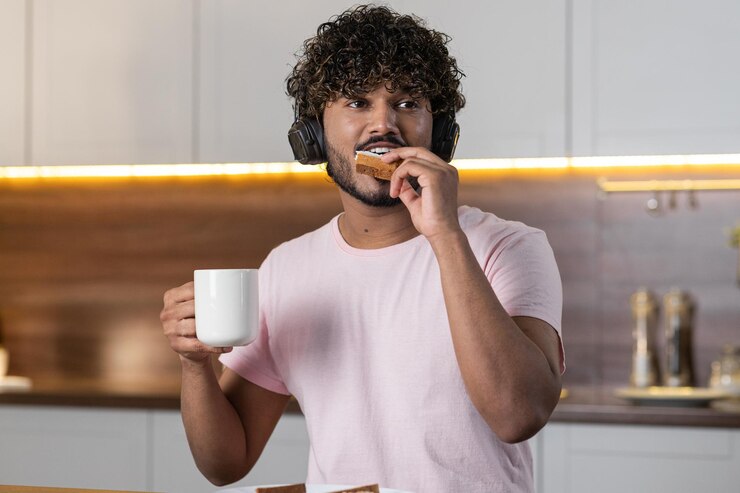 Foto um tipo mestiço positivo está na cozinha com uma xícara de chá ou café na mão e come uma sanduíche.