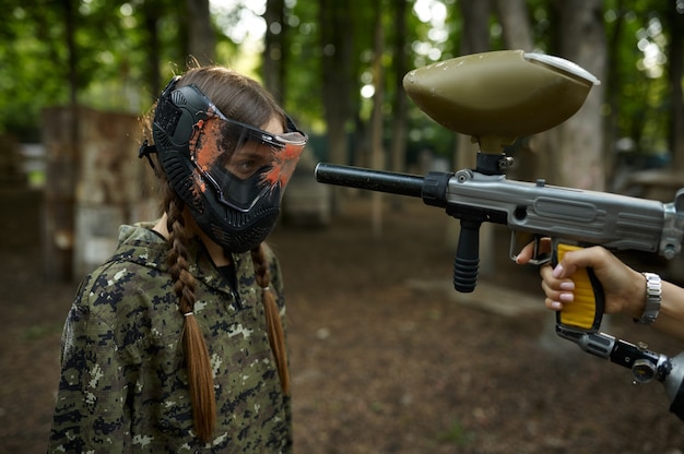 Um tiro de uma arma de paintball na cara da garota na máscara, parque infantil na floresta no fundo. Esporte radical com arma pneumática e balas ou marcadores de tinta, jogo de equipe militar ao ar livre