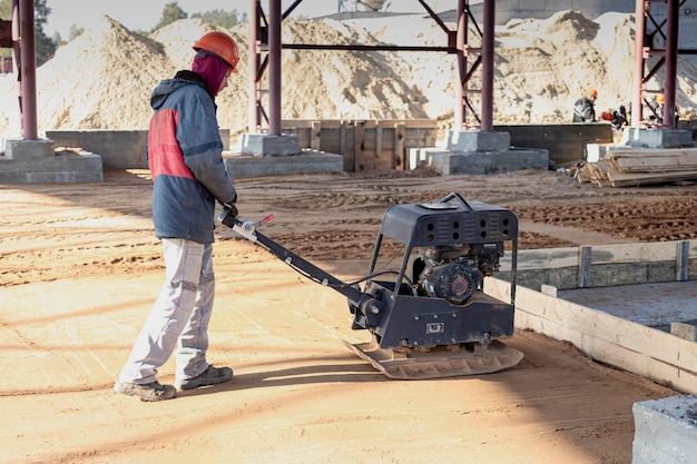 Um trabalhador compacta a terra com um compactador vibratório em um canteiro de obras em um dia ensolarado. Preparação da superfície para instalação de piso de concreto.