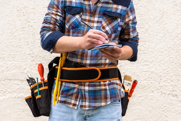 Um trabalhador da construção civil com uma camisa xadrez azul e ferramentas no cinto faz uma anotação com um lápis em um caderno.