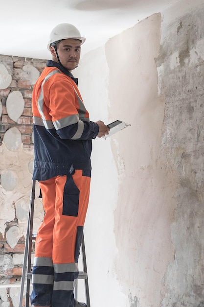 Um trabalhador da construção civil em um capacete branco e um uniforme de trabalho está de pé em uma escada Um homem está fazendo reparos em casa O conceito de consertar um apartamento ou casa