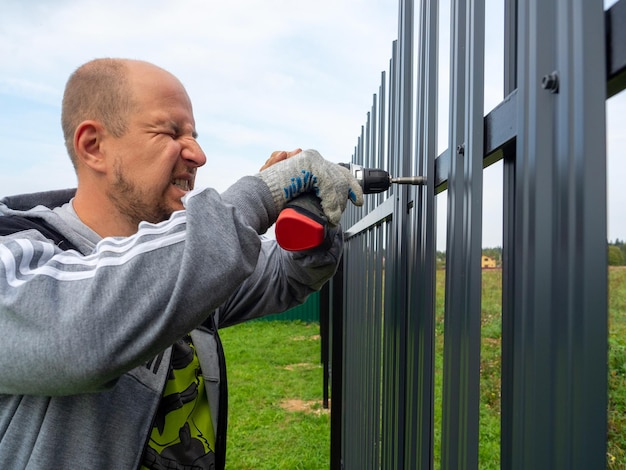 Foto um trabalhador do sexo masculino usando uma chave de fenda elétrica torce uma cerca de metal em cima da cerca construtor profissional trabalhador cercando território privado de segurança