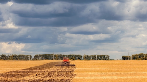 Um trator moderno laranja ara a terra em um campo de trigo dourado em um dia de verão, no céu uma nuvem cumulus