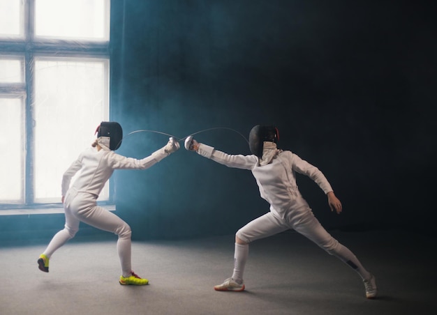 Um treinamento de esgrima no estúdio duas mulheres em trajes de proteção tendo um duelo cutucando com espadas uma na outra