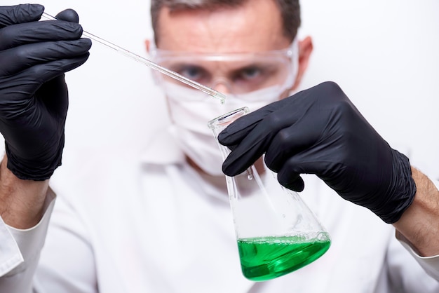 Um tubo de ensaio com uma gota e uma garrafa com um líquido verde nas mãos de um cientista nos óculos de proteção, uma máscara e luvas pretas.