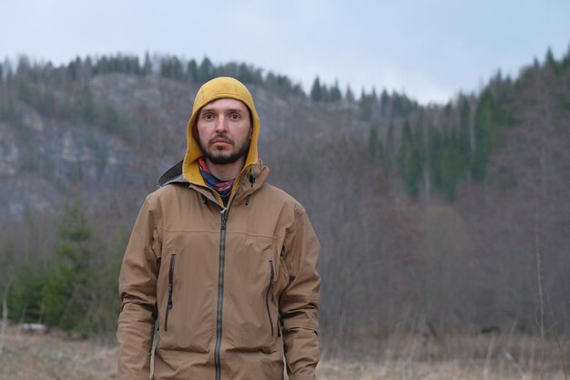 Um turista com uma jaqueta marrom e um capuz amarelo está contra o pano de fundo de montanhas arborizadas verdes