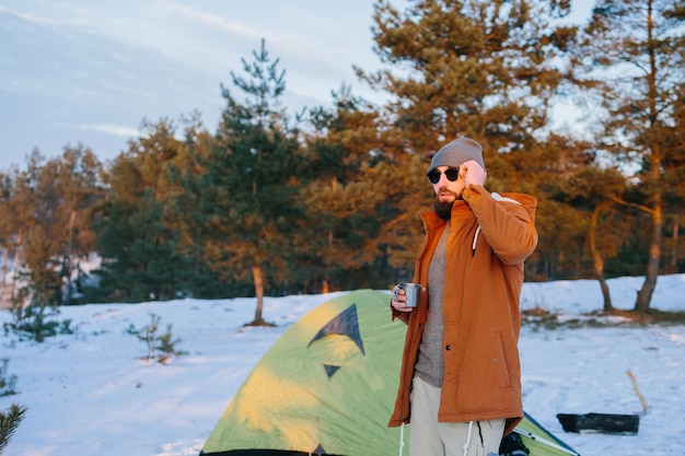 Um turista parado na tenda com uma xícara de chá ou café aprecia o pôr do sol na floresta de inverno