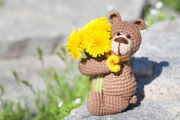 Um urso marrom feito malha pequeno com um lenço amarelo no jardim do verão. Brinquedo de malha, feito à mão, amigurumi