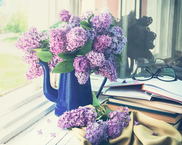 Um vaso azul de lilás fica no parapeito de uma janela ao lado de uma pilha de livros