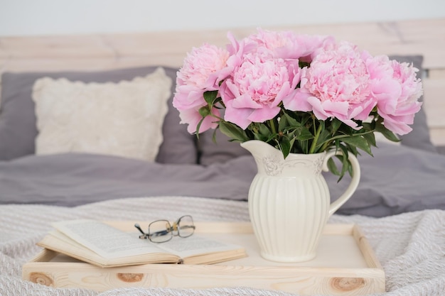 Um vaso com um buquê de peônias rosa um livro aberto e óculos em uma bandeja na cama