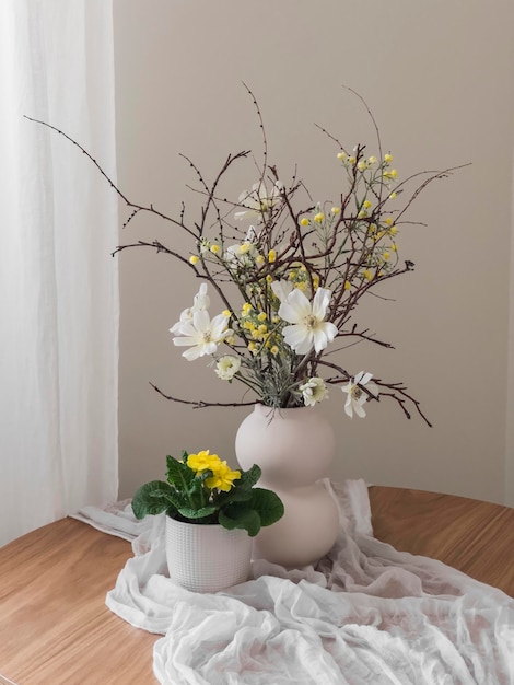 Um vaso de flores e uma flor de prímula caseira em uma panela em uma mesa redonda de madeira com uma toalha de mesa branca