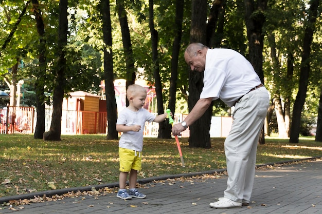 Um velho avô brinca com seu neto no fim de semana em um parque de diversões e alegremente sopra bolhas de sabão Um idoso, um aposentado e um garotinho passam tempo juntos