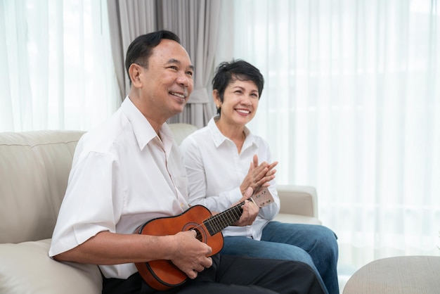 Um velho casal asiático canta uma canção com emoção feliz e toca ukulele juntos na sala de estar em sua casa