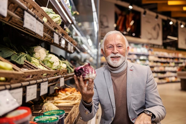 Um velho feliz no supermercado está comprando legumes frescos enquanto sorri para a câmera