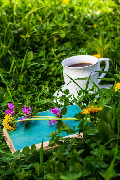 Um velho livro verde com flores silvestres e uma xícara de café em um prado verde em uma manhã de verão ou primavera