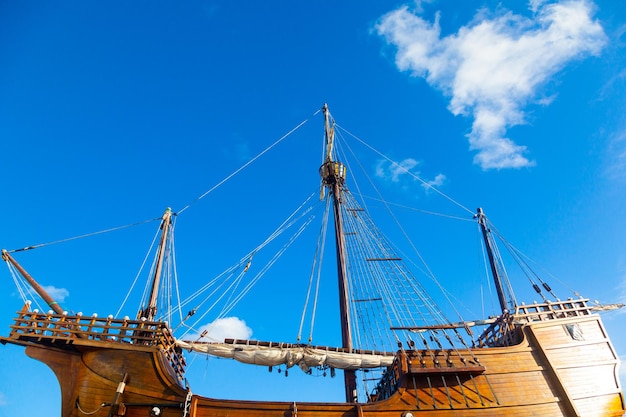 Um velho navio de caravela de madeira com velas dobradas contra um céu azul com nuvens brancas