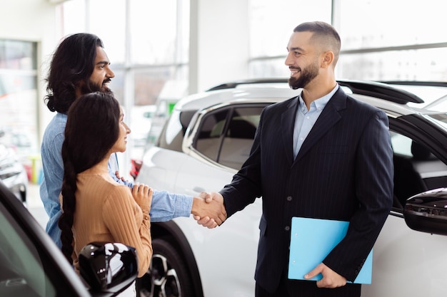 Um vendedor de carros em um terno está estendendo um aperto de mão para um homem indiano sorridente com uma mulher de pé